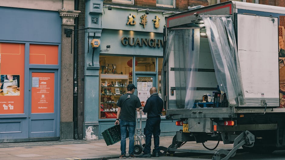 Inside a retail market space, a temporary setup features various wooden and wicker baskets, some open and containing produce or packaging materials. Several cardboard boxes with fresh produce, such as